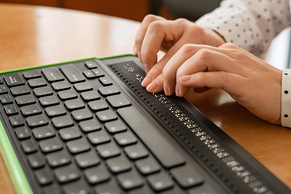 Nahaufnahme einer Person, die eine Braille-Tastatur mit eingebautem Braille-Display nutzt. Die Hände ertasten die erhabenen Punkte auf dem Display, während daneben eine herkömmliche Computertastatur liegt. Die Szene zeigt barrierefreie Technologie für sehbehinderte oder blinde Nutzer.